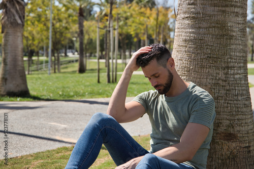 Young and handsome man, with blue eyes, beard, green t-shirt and jeans, sitting and leaning on the trunk of a palm tree, sad and lonely. Concept beauty, fashion, trend, sadness, loneliness.