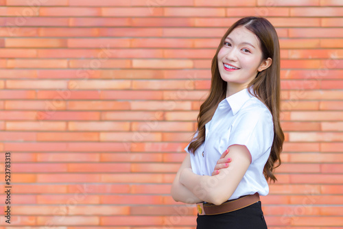 Portrait of an adult Thai student in university student uniform. Asian beautiful girl standing with her arms crossed on a brick background.