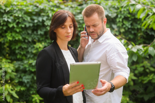 business partners man and woman are discussing something using smartphone tablet laptop. a team of two professional people discusses, decides, consults against the backdrop of green plants.