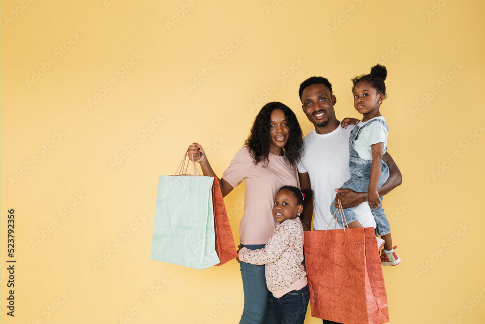 Young happy fun parents mom dad with two child kids daughters girls in casual clothes holding package bags with purchases after shopping isolated on yellow background. Young happy fun parents mom dad with two child kids daughters girls in casual clothes holding package bags with purchases after shopping isolated on yellow background.