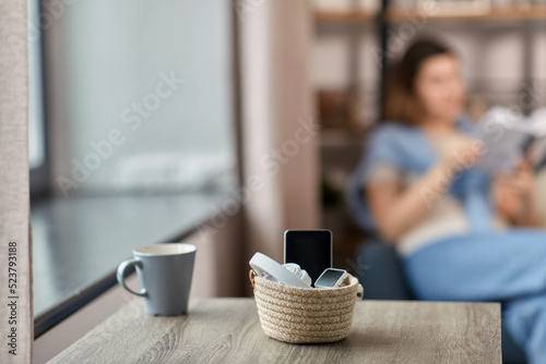 digital detox and leisure concept - close up of gadgets in basket on table and woman reading book at home