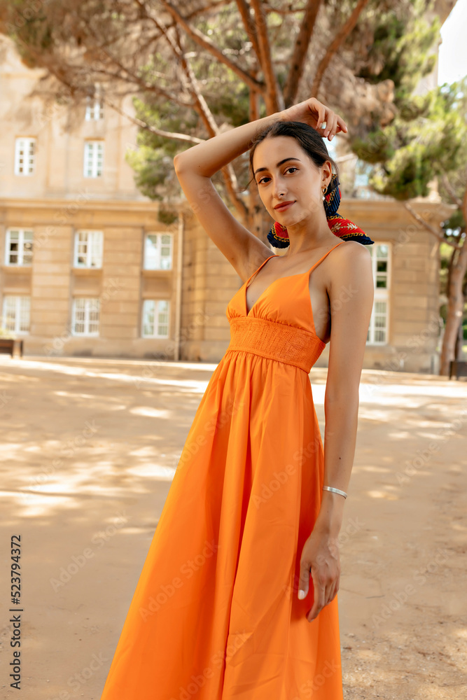Slim attractive stylish girl with dark hair and tanned skin wearing orange dress posing outdoor on city park background in sunny warm day