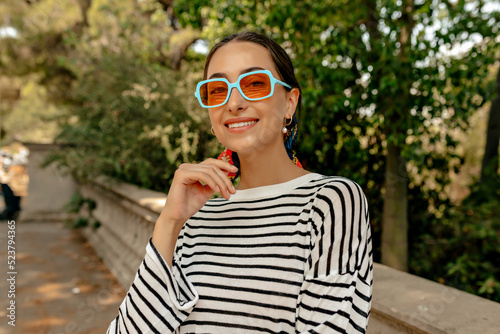 Happy charming cute lady with wonderful smile wearing bright summer sunglasses and striped shirt looking at camera with wonderful smile and walking on street in summer warm day