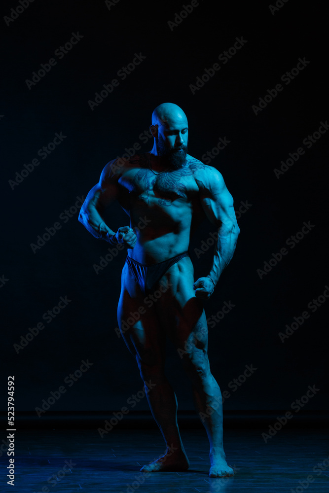 Athletic man demonstrates muscles in the light of a blue light filter on a dark background.