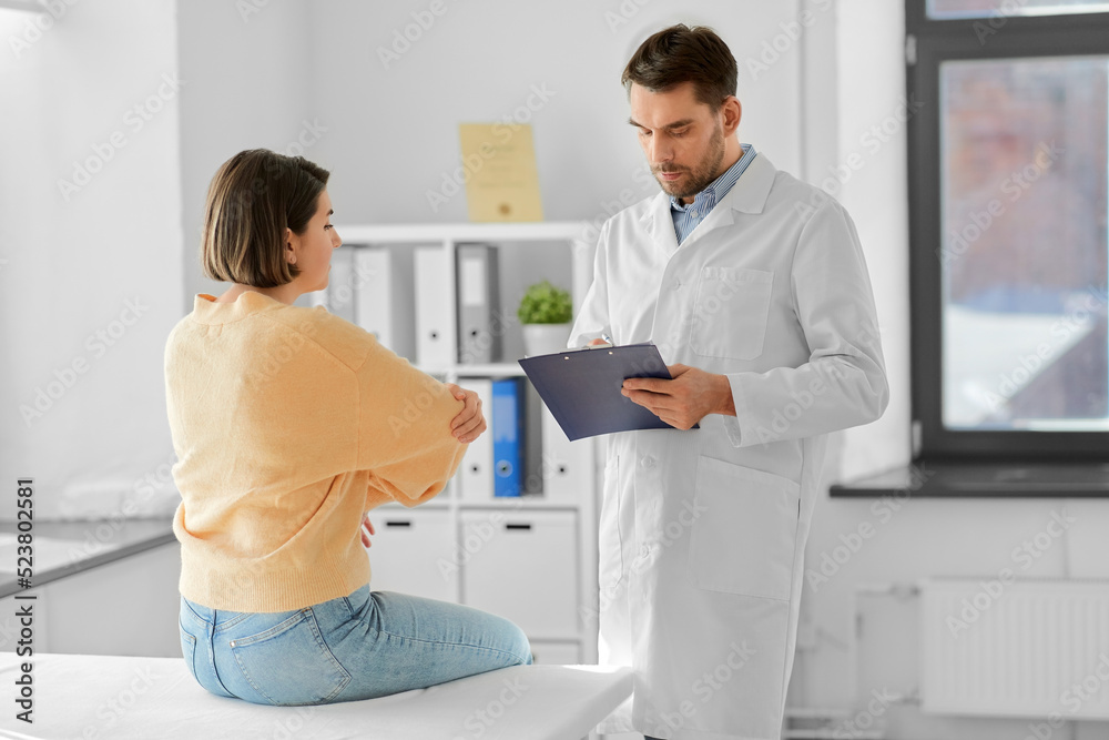 Obraz premium medicine, healthcare and people concept - male doctor with clipboard talking to woman showing her sore arm patient at hospital