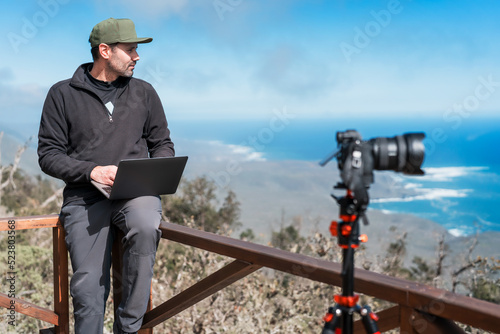 mature person with a laptop and photographic camera sitting on lookout point working over the pacific ocean portrait  photo