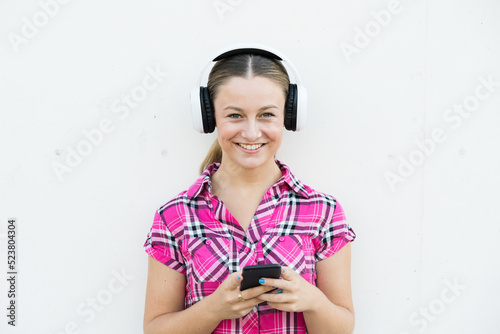 Pretty blonde teenager girl wearing headphones and using phone. Young woman listening music or podcast against concrete wall.