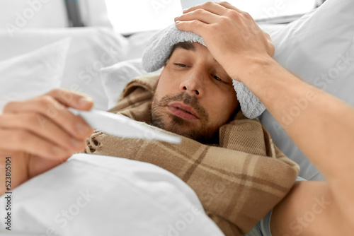 people, health and fever concept - sick man with cold compress on his forehead measuring temperature by thermometer lying in bed at home