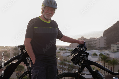 Fototapeta Naklejka Na Ścianę i Meble -  Smiling senior sporty man with helmet standing on hill with electric bicycle, healthy activity for retiree man enjoying vacation