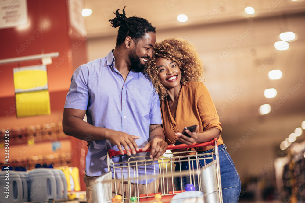 African American couple choosing products using phone during grocery shopping in modern supermarket