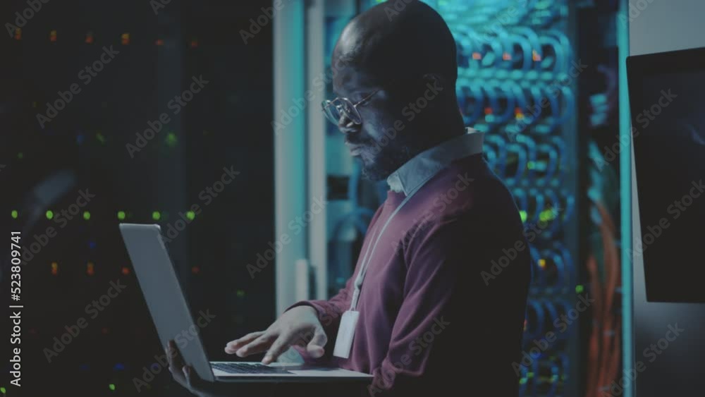 Side view of African American server administrator coding on laptop in dark server room of data center