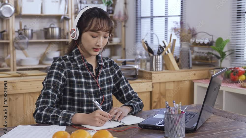 concentrated asian female is writing notes and looking at the computer screen while listening to lectures from earphones at home dining table.