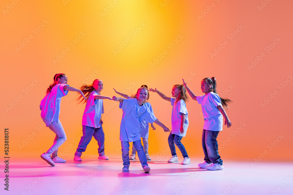 Dance group of happy, active little girls in t-shirts and jeans in action isolated on orange background in neon. Concept of music, fashion, art, childhood