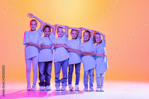 Portrait of cheerful, active little girls, happy kids dancing isolated on orange background in neon light. Concept of music, fashion, art, childhood, hobby