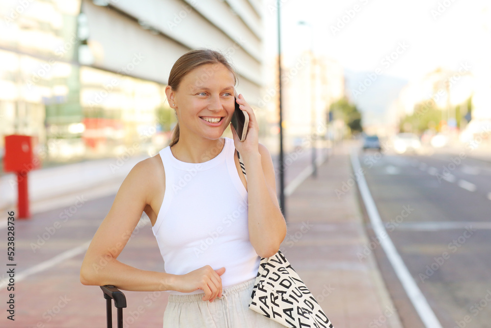 Beautiful stylish young woman is using smartphone, with suitcase outdoors by the airport or railway station waits for taxi cab. Attractive smiling girl ready to travel with luggage in the modern city. Beautiful stylish young woman is using smartphone, with suitcase outdoors by the airport or railway station waits for taxi cab. Attractive smiling girl ready to travel with luggage in the modern city.