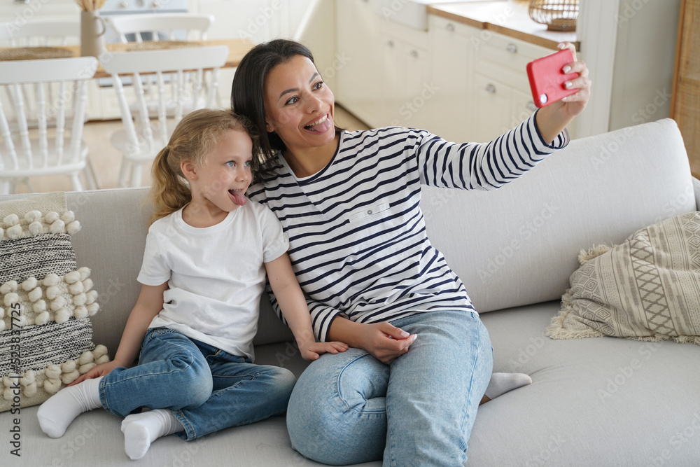 Little daughter, mother show tongues taking selfie of family with phone having fun together at home