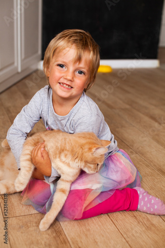 A girl with a red cat in her arms. A smiling blonde girl with short hair plays with a pet at home