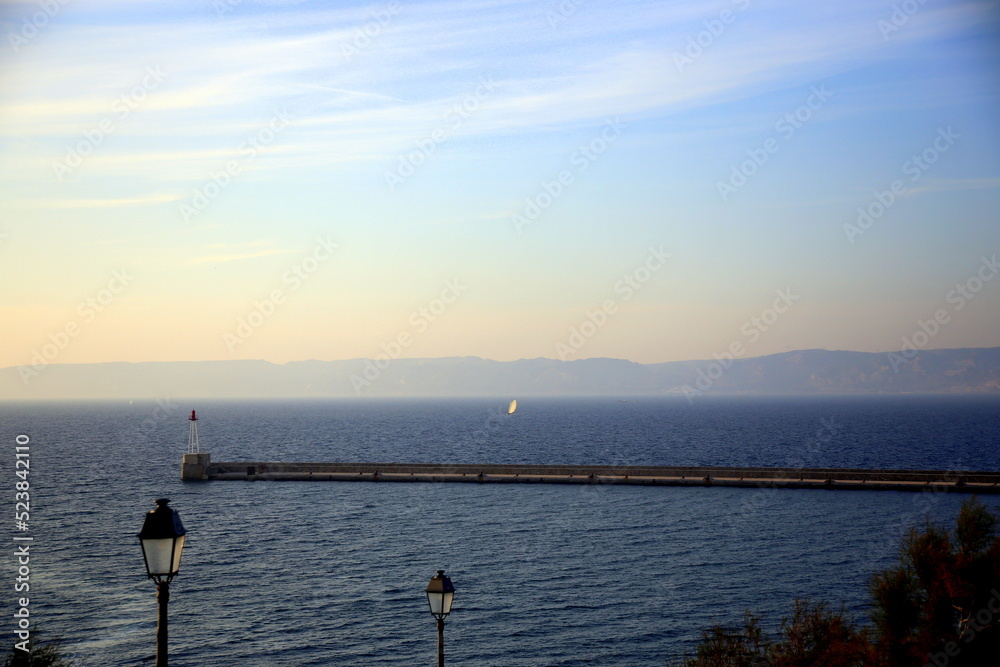 Seascape with two lampposts near the pier, a sailboat in the background against the coast, at sunset, Marseille, France
