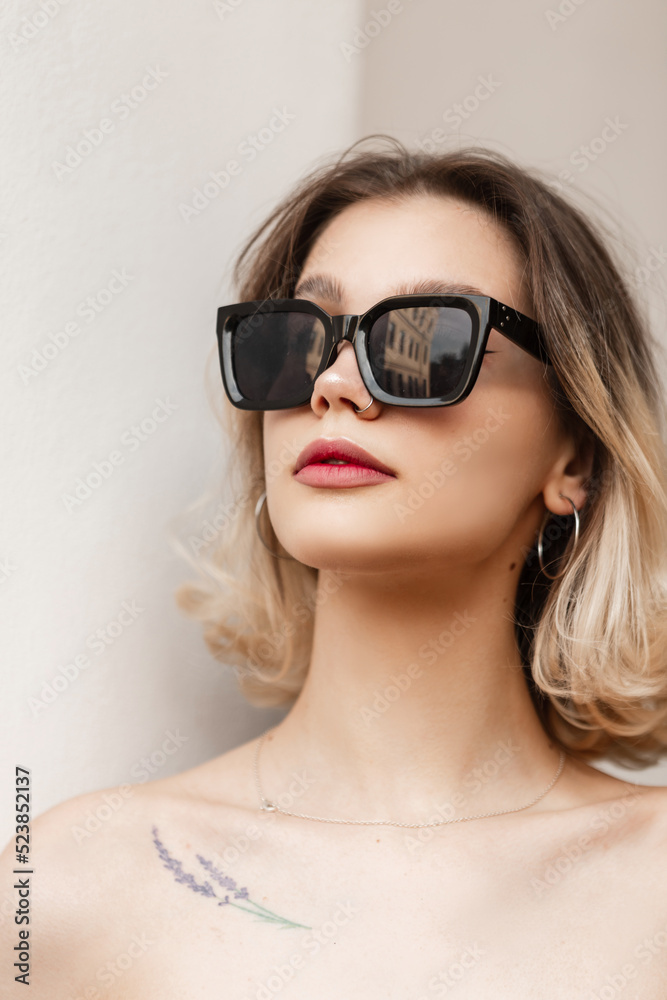 Beautiful female headshot portrait of a pretty stylish young girl with a nose piercing with black fashion sunglasses on a white background on the street Beautiful female headshot portrait of a pretty stylish young girl with a nose piercing with black fashion sunglasses on a white background on the street