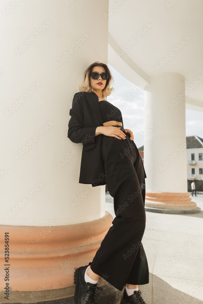 Elegant vogue beautiful young business woman model with cool trendy sunglasses in fashion strict black outfit with jacket, pants, top and shoes standing near a white vintage column in the city Elegant vogue beautiful young business woman model with cool trendy sunglasses in fashion strict black outfit with jacket, pants, top and shoes standing near a white vintage column in the city