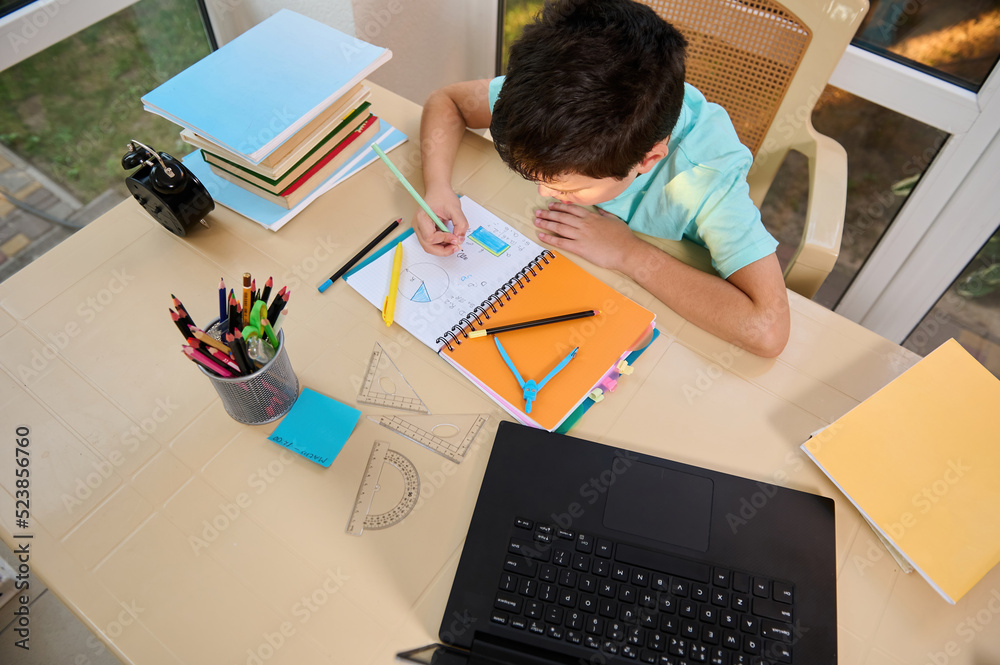 Top view. School boy solves maths and geometry examples, sitting at a table with school supplies, measuring rulers, stacked textbooks, black alarm clock and laptop, broadcasting an online video lesson Top view. School boy solves maths and geometry examples, sitting at a table with school supplies, measuring rulers, stacked textbooks, black alarm clock and laptop, broadcasting an online video lesson