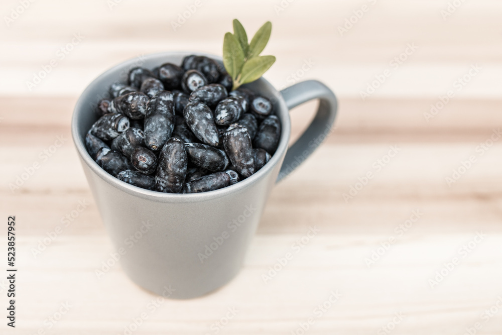 Kamchatka berry / haskap berry / honeysuckle (Lonicera caerulea) in a gray cup, decorated with green leaves, standing on a wooden board. The first summer fruit, perfect for desserts. Kamchatka berry / haskap berry / honeysuckle (Lonicera caerulea) in a gray cup, decorated with green leaves, standing on a wooden board. The first summer fruit, perfect for desserts.