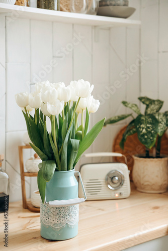 Bouquet of fresh white tulips in a decorative jar on a wooden kitchen worktop against a blurred background of retro radio and potted plants in a modern white scandinavian kitchen interior. photo