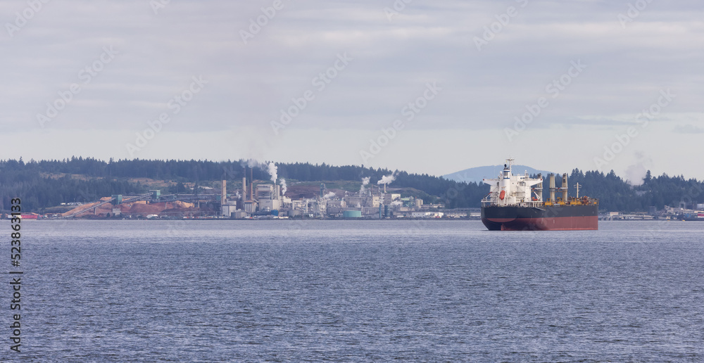 Industrial Processing Plant and Container Ship passing by on a cloudy day. Nanaimo, Vancouver Island, British Columbia, Canada.