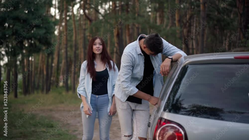Stressed handsome man leaning on broken car with woman jumping gesturing calling help. Front view of Caucasian boyfriend and girlfriend with automobile in forest. Trip problem Stressed handsome man leaning on broken car with woman jumping gesturing calling help. Front view of Caucasian boyfriend and girlfriend with automobile in forest. Trip problem