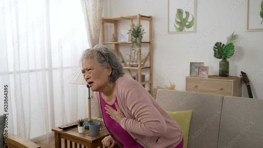 asian old female suffering sudden sharp pain from heart attack is moaning and clutching her chest in agony as she is sitting down on the sofa at home