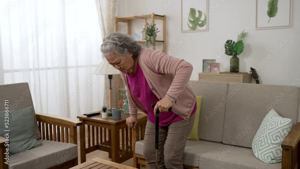 unwell asian grandmother moving around with cane walking stick at home is sitting down to rest on the couch and rubbing her painful back in agony at daytime in the living room unwell asian grandmother moving around with cane walking stick at home is sitting down to rest on the couch and rubbing her painful back in agony at daytime in the living room
