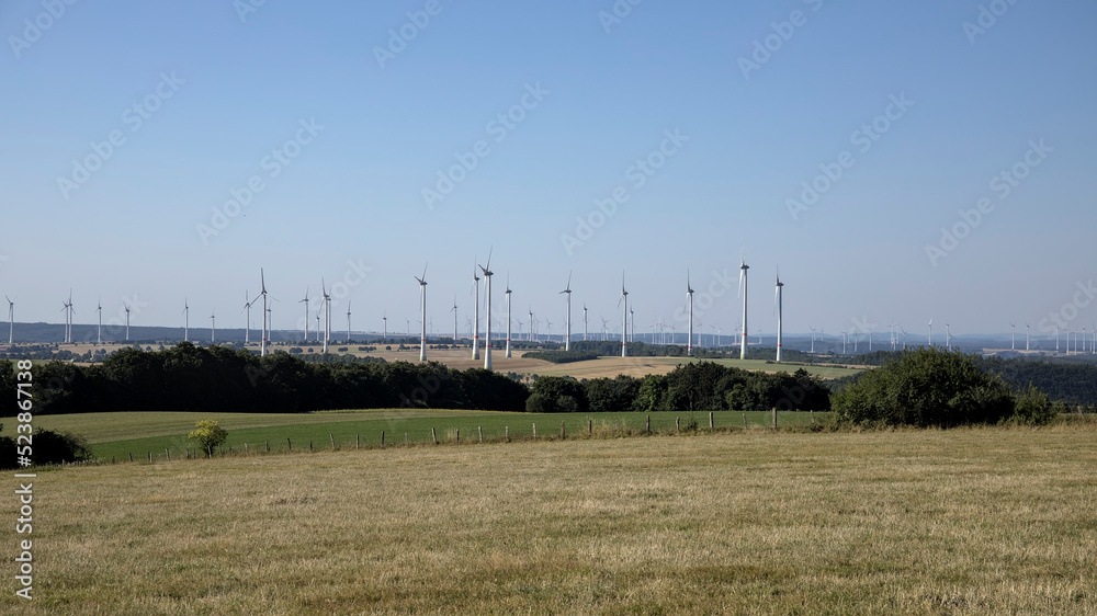 Many wind turbines in rural environment at the horizon with fields, bushes and forests Many wind turbines in rural environment at the horizon with fields, bushes and forests