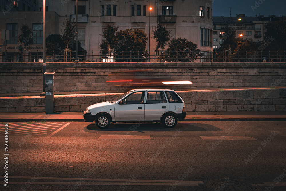 Classic European hatchback parked under street light with can speeding past behind it making motion blue, in Budapest, Hungary 