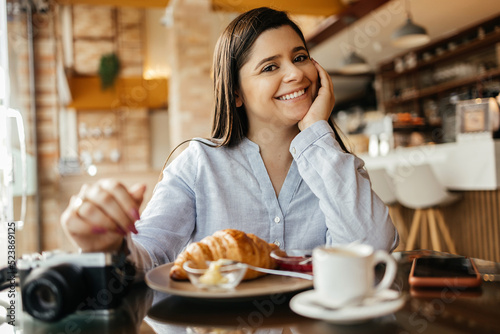 Smiling woman sitting at table and having breakfast in cafe with photo camera.