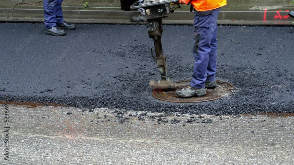 Replacing old asphalt when repairing the road surface. A road worker in uniform lays a manhole cover on a well on a road with new asphalt. Road repair in Europe. Reconstruction of the road surface Replacing old asphalt when repairing the road surface. A road worker in uniform lays a manhole cover on a well on a road with new asphalt. Road repair in Europe. Reconstruction of the road surface