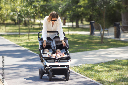 Lifestyle young casual caucasian mother with two little twin girls in a double baby stroller walks in city the park  photo