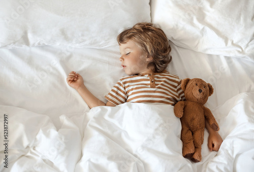 Top view of a cute little baby hugging his toy teddy bear and sweet sleeping under a blanket on a soft comfortable pillow, white clean bedding in cozy bed at home
