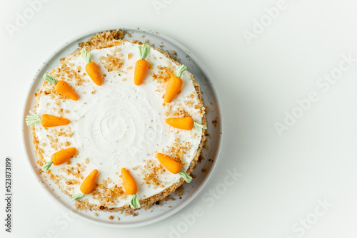 Delicious carrot cake decorated with mastic sweet carrots. Homemade carrot cake with yellow crumbs in the white plate on the white background. Flat lay