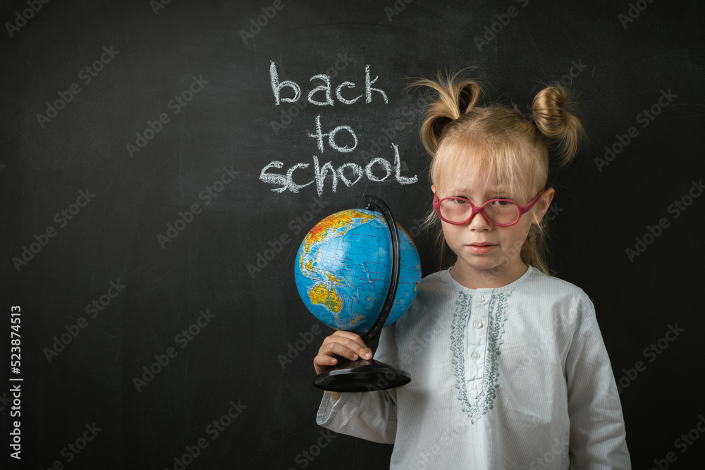 Fototapeta premium Portrait of little caucasian girl wearing glasses. Elementary school student holding the globe on the black school board background. Back to school concept