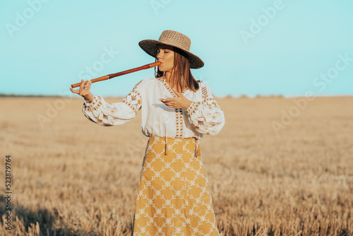 Woman playing on woodwind wooden flute - ukrainian telenka or tylynka in wheat field. Folk music concept. Musical instrument. Musician in traditional embroidered shirt - Vyshyvanka. photo