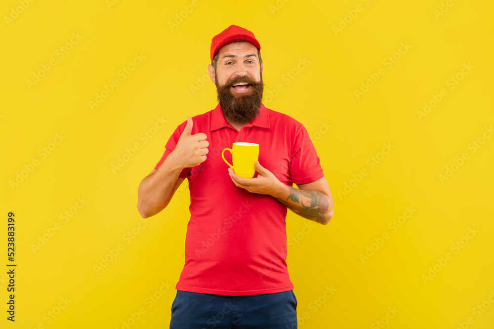 Naklejka premium Happy bearded man in casual red cap and tshirt giving thumb holding mug yellow background, coffee
