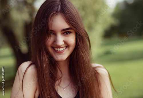 Beautiful toothy smiling teen woman looking happy outdoors on summer green trees background. Closeup portrait in bright sunny day.