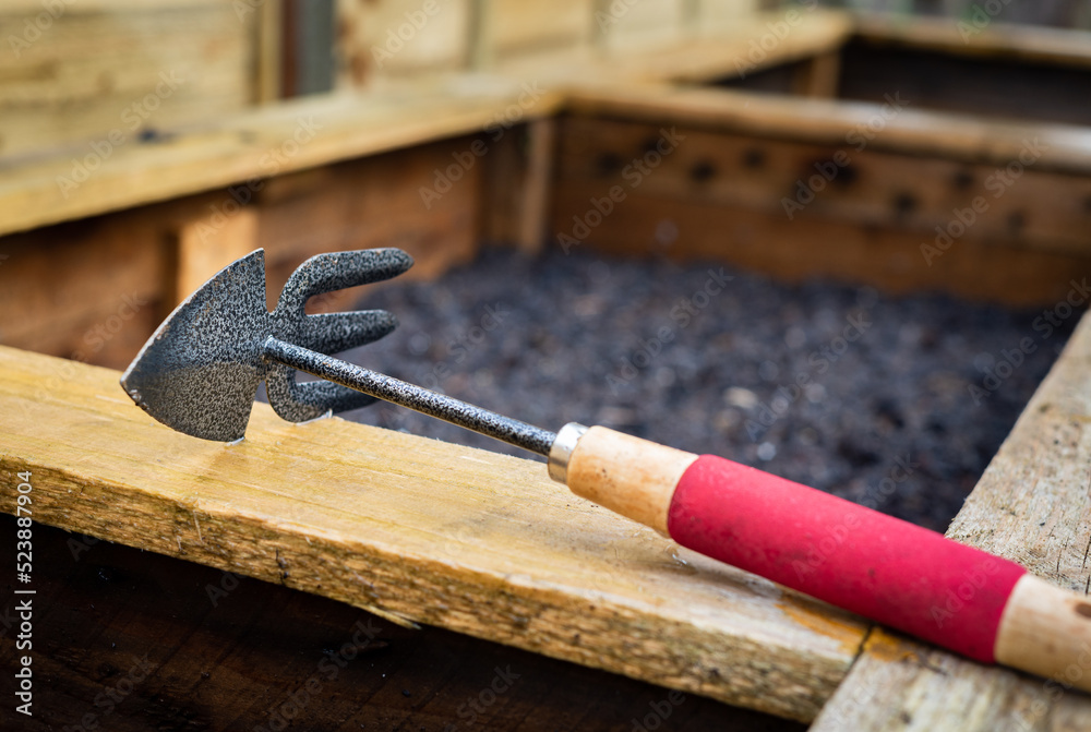 Double sided hoe and rake garden hand tool on top of freshly planted wooden vegetable planter box. Double sided hoe and rake garden hand tool on top of freshly planted wooden vegetable planter box.