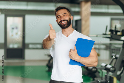Portrait of personal trainer with clipboard showing thumb up at gym