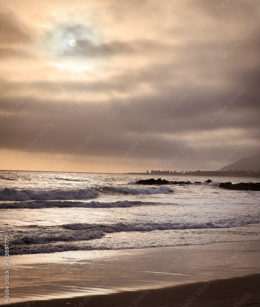 Moody Sunset with waves crashing.  Sunbeams streaming through the clouds with mountains in the background.