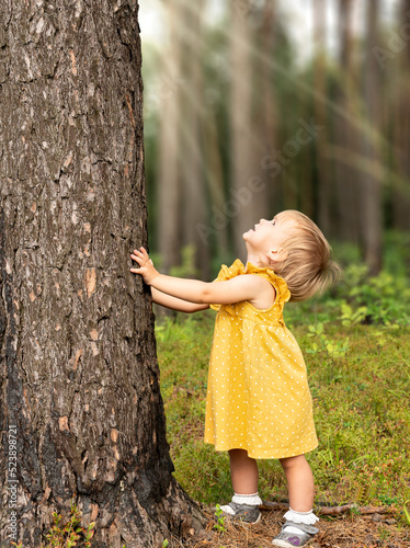A little girl in a yellow dress leaned her hands against a tree trunk and looks up