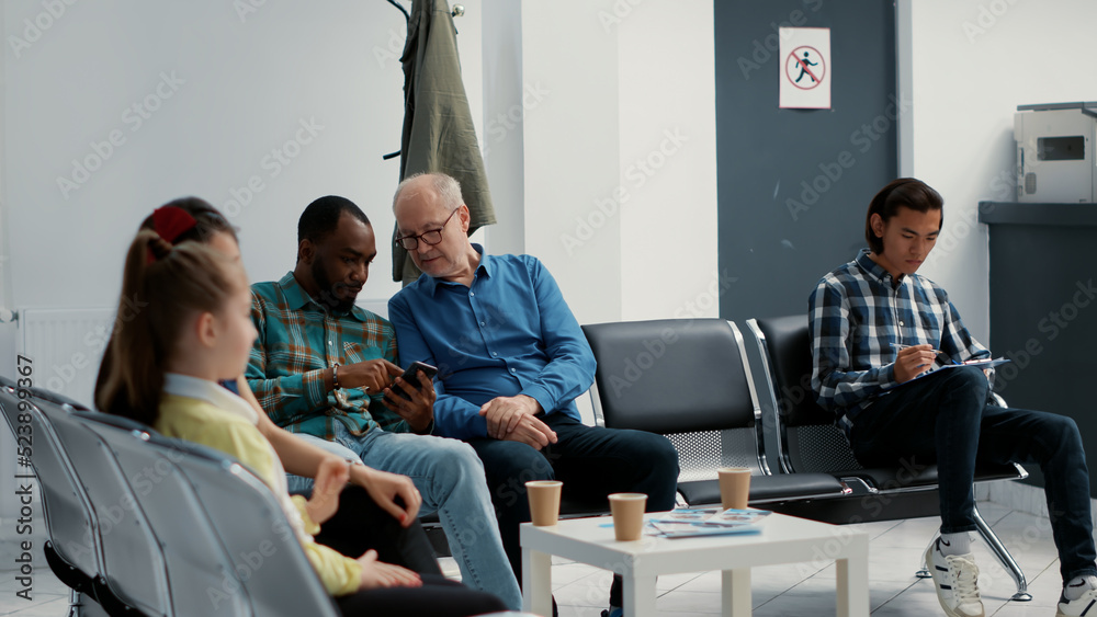 African american man and senior patient waiting together at hospital reception, looking at online website on smartphone. Diverse people sitting in waiting room lobby at medical clinic. African american man and senior patient waiting together at hospital reception, looking at online website on smartphone. Diverse people sitting in waiting room lobby at medical clinic.