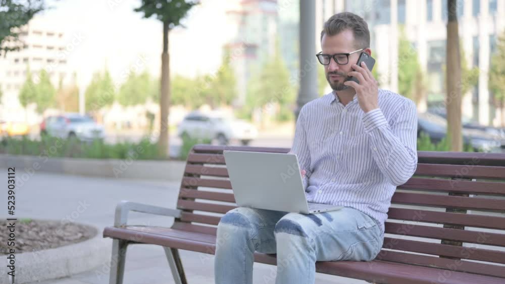 Young Adult Man Talking on Phone and using Laptop while Sitting Outdoor on Bench Young Adult Man Talking on Phone and using Laptop while Sitting Outdoor on Bench