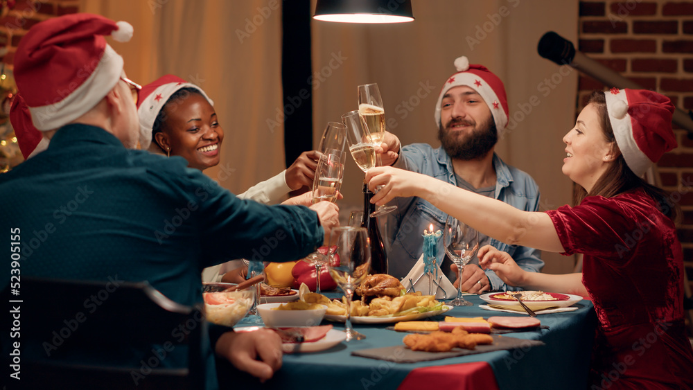 Festive diverse people wearing traditional hats while clinking champagne glasses at Christmas dinner. Happy family celebrating winter holiday with home cooked food and sparkling wine. Handheld shot Festive diverse people wearing traditional hats while clinking champagne glasses at Christmas dinner. Happy family celebrating winter holiday with home cooked food and sparkling wine. Handheld shot