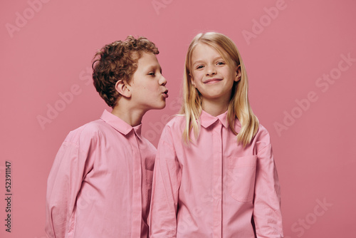 cute, beautiful children are standing in pink clothes on a pink background and the boy reaches out to kiss the girl on the cheek. The topic of relationships
